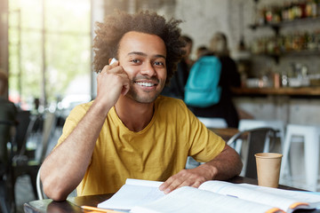 Obraz premium Smiling black youngster sitting in cafeteria speaking over smart phone having broad smile and good mood while having rest for minute after studying with books. Delightful dark-skinned student