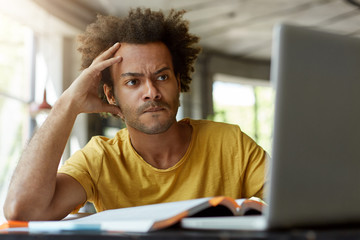 Attentive black young male sitting indoors in front of opened laptop being very serious while reading scientific article online trying to find main point of it and to write review on this topic