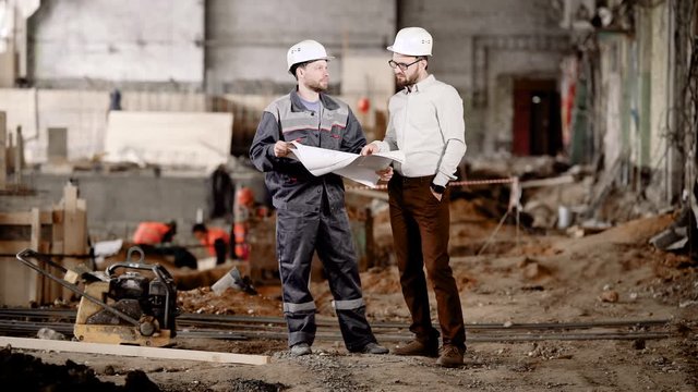 Two men in safety helmets standing in construction area and looking at scheme of future building. Foreman pointing to drawing and explaining to the worker highlights of current assembling.