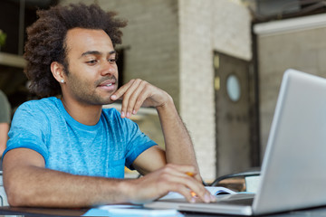 People, modern technology, education, learning and knowledge concept. Handsome comfident young Afro American man sitting in front of open laptop, studying, taking online course on modern art