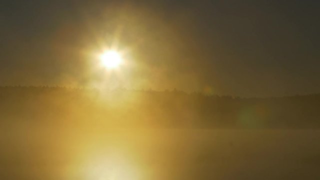 Summer sunset on forest lake. View on Lake with bright Sun over distant cape and evening glow reflecting in calm water at midnight sun. Fog on the river in the moonlit night