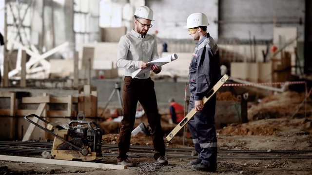 Two men in white safety hardhats standing in building site. Engineer bringing contsruction plan and showing it to worker. House-builder holding spirit level and investigating draft with his colleague.