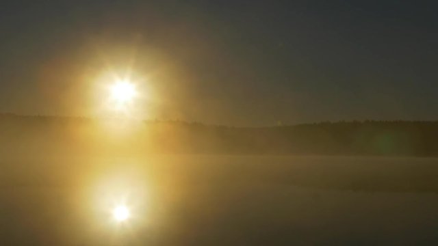 Summer sunset on forest lake. View on Lake with bright Sun over distant cape and evening glow reflecting in calm water at midnight sun. Fog on the river in the moonlit night