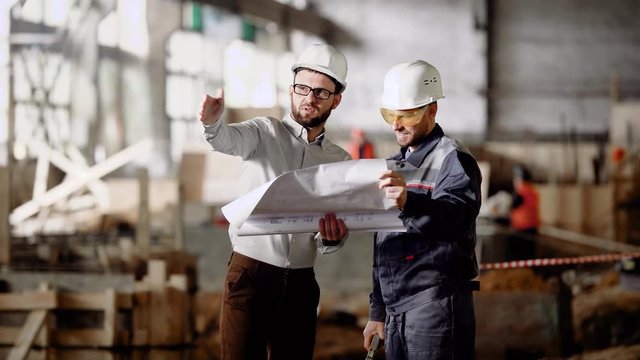 Two engineers standing in building site and holding construction plan. Men in white hardhats working discussing detailes of design project. Foreman explaining to worker modification of installation.