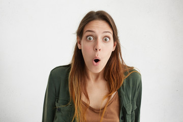 Studio shot of amazed woman with bugged eyes and opened mouth staring in camera after hearing latest news on television. Surprised woman with widely opened eyes and mouth posing over white wall