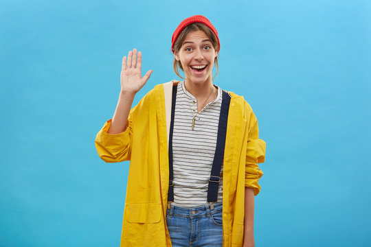 Happy positive young female dressed casually waving with her palm greeting friends, showing friendly sign isolated over blue background. Smiling pleasant-looking woman raising her hand with pleasure