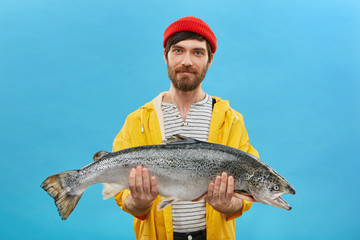 Bearded fisherman in yellow anorak and red hat holding huge fish in hands, demonstrating his...