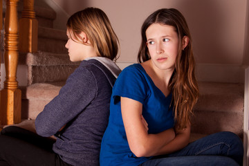 Two teenage girls sitting on the stairs back to back looking angry with each other.