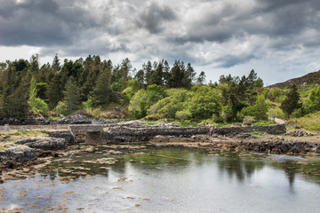 Fototapeta na wymiar Assynt Peninsula, Scotland - June 7, 2012: Short stone bridge over creek landing into Atlantic Ocean inlet South of Loch An Arbhair under heavy dark sky. Forested green hills,ocean waters, road.