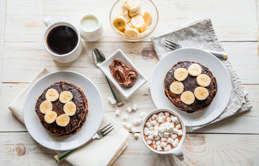 Breakfast for couple included oat pancakes with banana, nutella, honey, with coffe and milk and cacao with marshmallow on white wooden  background