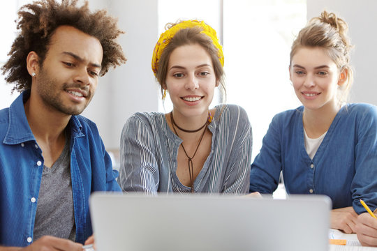 Three mixed race people watching films at laptop using free internet connection having happy expressions. Friends of different races calling their best friend abroad over computer using wi-fi
