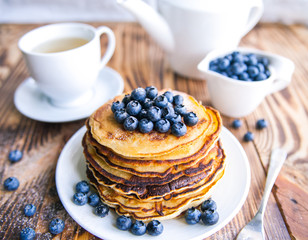 Pancakes healthy breakfast with blueberries, bog whortleberry, cup of green tea, cup of blueberries and teapot on brown wooden background 