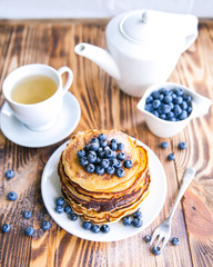 Pancakes healthy breakfast with blueberries, bog whortleberry, cup of green tea, cup of blueberries and teapot on brown wooden background 