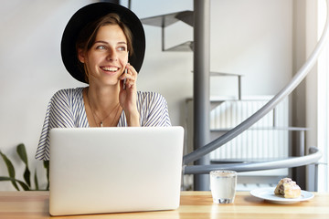 Elegant woman dressed in hat and formal blouse having conversation over smart phone while sitting at her workplace with laptop and lunch looking aside with pleasant smile noticing someone in office