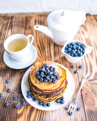 Pancakes healthy breakfast with blueberries, bog whortleberry, cup of green tea, cup of blueberries and teapot on brown wooden background 