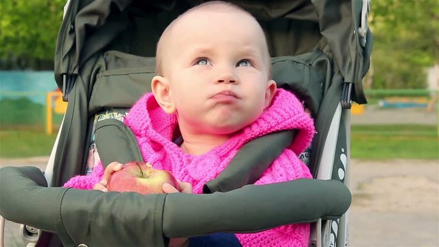 Baby girl sits in a baby stroller and eats an apple