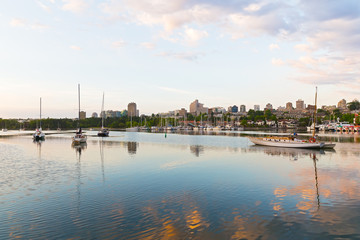 Obraz premium Yachts on the water in early morning and urban landscape on horizon. Sunrise touched water surface of False Creek in Vancouver, Canada.