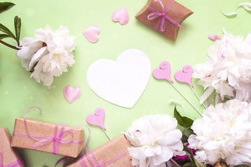 White peonies with empty heart-card, gift boxes and decorative hearts on a green background. Copy space.