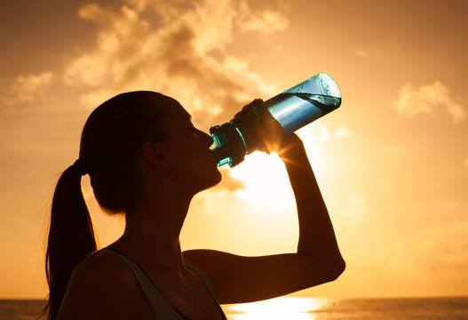 Young woman drinking bottle of water against a sunset. Fitness and health.