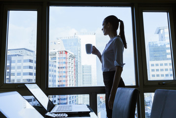 Woman standing next to office window drinking a cup of coffee. 