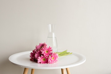 Pink gerberas lying on small white round table next to glass vase against neutral wall background (selective focus)