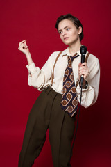 Fashionable beautiful singer girl in retro coat, tie and pants with microphone in hands posing on red background.