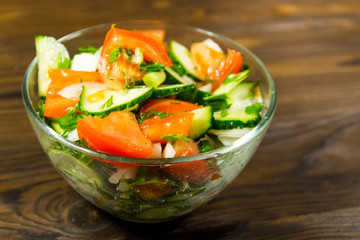 Fresh salad with tomato, cucumber, onion, parsley and dill in glass bowl on wooden table