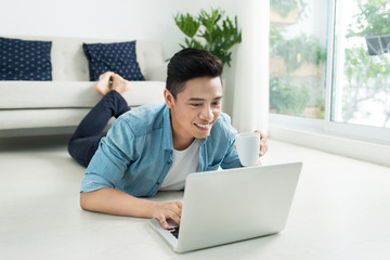 Asian man lying on the floor with laptop and coffee.