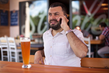 Young  bearded man talking on phone and drinking beer in bar