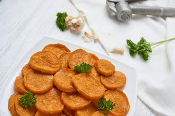 Baked sweet potatoes in white plate on a white wooden table with parsley, garlic and salt. White wooden rustic picnic table scenery from above.