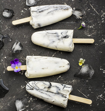 Yoghurt refreshing marble ice cream popsicle in trendy fashion colors on a dark background with edible flowers of garden violas. Selective focus. Top View