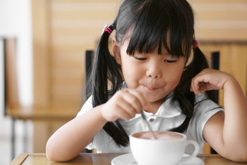 Asian children cute or kid girl smile and delicious enjoying with hot cocoa or chocolate drinking in white cup for breakfast and holding spoon for stir and scoop at morning on table in home or cafe