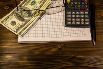Notebook with dollars, pen, glasses and calculator on wooden desk
