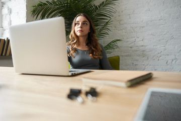 Indoor shot of serious beautiful female student with long hair sitting at table in front of open laptop computer, doing homework, looking for information on Internet. Selective focus on girl's friend