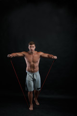 Fototapeta na wymiar Full lenght portrait of shirtless young European bodybuilder exercising in gym, building bicep and tricep using resistance rubber band, wearing shorts. Physical training, fitness and sports concept