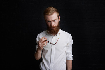 Indoor shot of good-looking confident successful young businessman in formal white shirt standing isolated against studio wall background, holding sunglasses, having serious and concentrated look