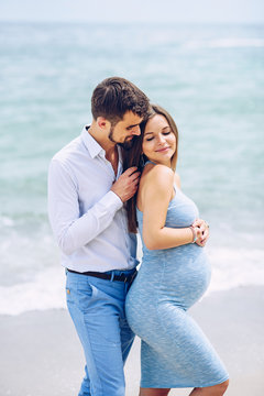 Courageous and handsome man in a white shirt and blue trousers  hugs her beautiful pregnant wife in a blue dress against the background of the sea.