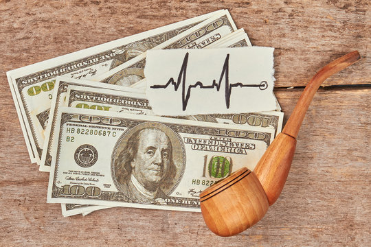 Cost of tobacco cigarette addiction. Dollars, tobacco pipe, image of heartbeat, wooden background.
