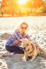 Portrait of young beautiful woman in sunglasses sitting on sand beach with golden retriever dog. Girl with dog by sea. Sun flare