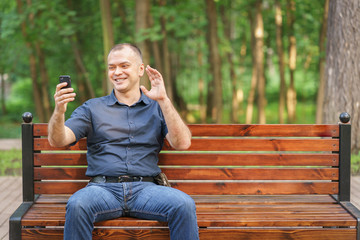 adult man around 35-40 with smile is talking on video communication by phone at summer park