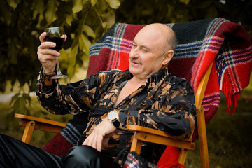 Obraz premium Mature stylish senior in his garden sit in red plaid armchair and tasting red wine