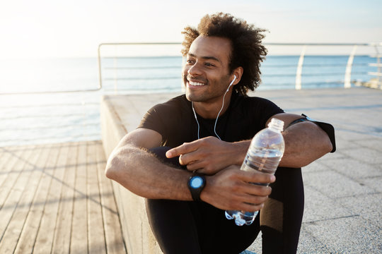 Cheerful dark-skinned muscular athlete in black sport clothing sitting on pier after sport activities wearing white earphones. Smiling runner enjoying morning workout and drinking water. Motivation