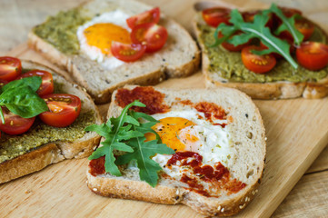 Fototapeta na wymiar Toasts with egg, pesto and tomato sauce, arugula, spinach, cherry tomato and multi cereal bread on wooden background. Healthy breakfast. Balanced meal