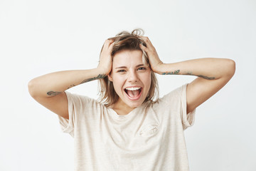 Emotional cheerful young tattooed girl smiling with opened mouth looking at camera touching hair over white background.