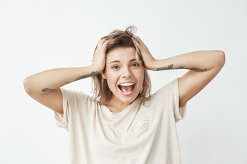 Emotional cheerful young tattooed girl smiling with opened mouth looking at camera touching hair over white background.