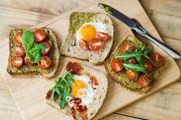 Breakfast toasts on wooden cutting board. Healthy sandwiches with cherry tomato, egg, spinach, arugula, pesto sauce, tomato sauce and bread with sesame seeds. Balanced breakfast