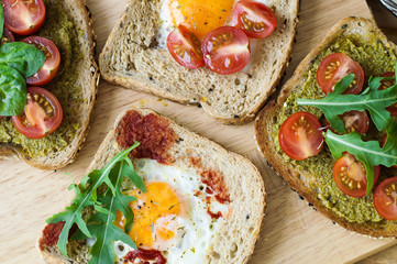 Toasts with healthy bread with sesame seeds, egg, cherry tomato, arugula, spinach, pesto and tomato sauce on wooden board. Breakfast meal
