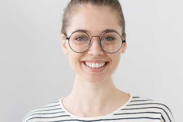 Closeup of young European teenage girl isolated on grey background wearing eyeglasses with thin black rim, smiling to photographer, looking happy, positive and optimistic, open for communication.