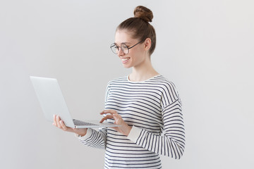Indoor photo of positive teenage girl isolated on grey background standing with laptop, smiling at...