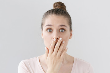 Indoor shot of beautiful teenage girl isolated on gray background dressed in light pink top, covering mouth with hand in sign of deep amazement and shock, hardly believing news she is receiving.